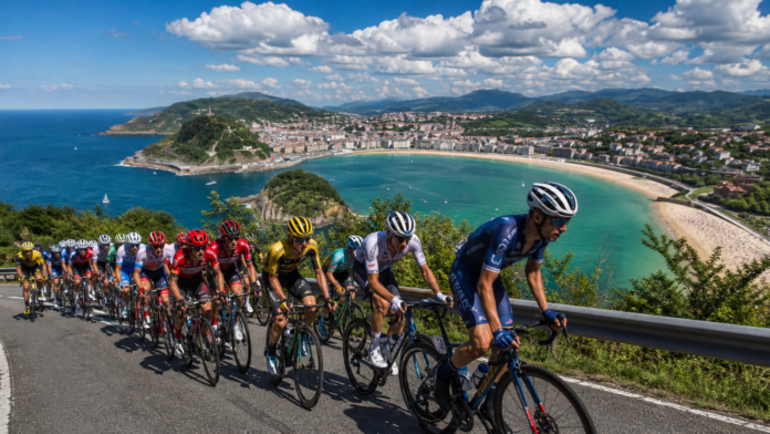 clasica-san-sebastian-capa-pelotao-costa-basca Pelotão profissional da Clásica San Sebastián pedalando pela costa do País Basco com a praia de La Concha e a baía de San Sebastián ao fundo em dia ensolarado de verão