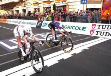 Milão-Sanremo 2026: Pogacar vence! adej Pogacar sprintando contra Tom Pidcock na Via Roma em Sanremo vencendo a Milão-Sanremo 2026, jersey branca, vermelha e preta da UAE Team Emirates-XRG com listras arco-íris de campeão mundial, manga esquerda rasgada e suja da queda, palmeiras e edifícios elegantes ao fundo, Mar Mediterrâneo ao longe, céu nublado dramático, asfalto molhado