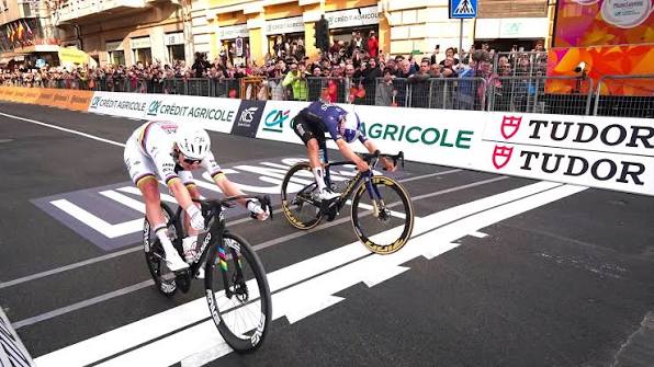 pogacar-vence-milao-sanremo-2026-capa adej Pogacar sprintando contra Tom Pidcock na Via Roma em Sanremo vencendo a Milão-Sanremo 2026, jersey branca, vermelha e preta da UAE Team Emirates-XRG com listras arco-íris de campeão mundial, manga esquerda rasgada e suja da queda, palmeiras e edifícios elegantes ao fundo, Mar Mediterrâneo ao longe, céu nublado dramático, asfalto molhado