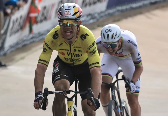 wout-van-aert-vence-paris-roubaix-2026-capa Wout van Aert cruza a linha de chegada no Vélodrome André-Pétrieux vencendo o Paris-Roubaix 2026, braço direito erguido com um dedo apontado para o céu em homenagem a Michael Goolaerts, velódromo ao ar livre de pista de concreto cinza com arquibancadas simples e chaminé industrial ao fundo, céu nublado, bandeiras belgas na arquibancada